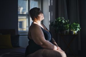 Depressed lonely fat woman sitting and looking through the window at home